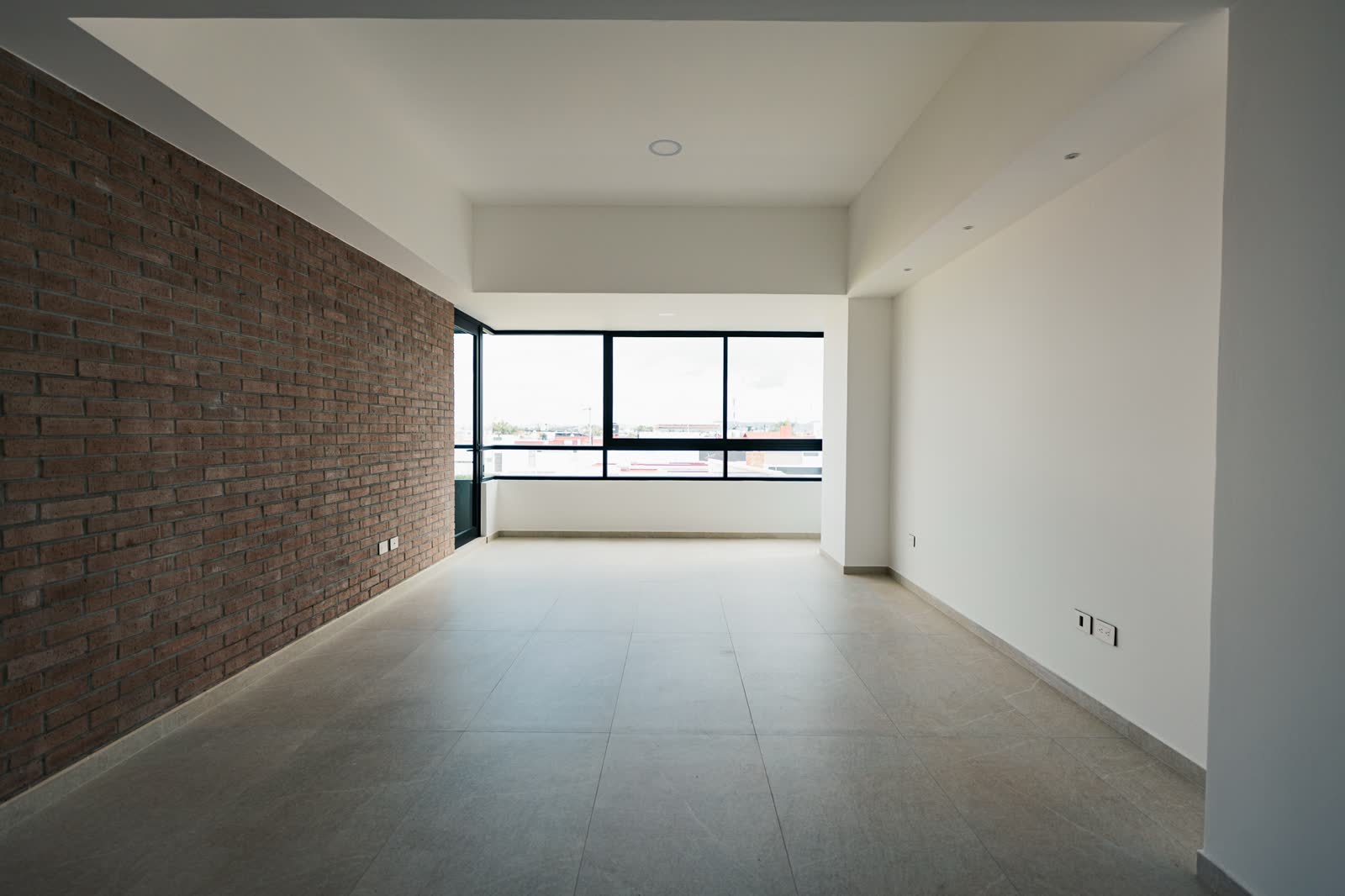 Sala vacía con ventanal de piso a techo, muro de ladrillo y luz natural